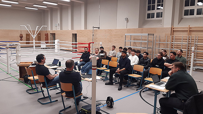 Ein Blick auf die Abteilungsversammlung. Mitglieder sitzen auf Stühlen und hören dem Vortrag der Abteilungsleitung zu. Im Hintergrund ist die frisch sanierte Halle erkennbar mit aufgebautem Boxriung und Sandsäcken.