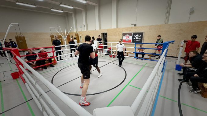 Zwei Boxer im Ring, die einen Sparringskampf durchführen. In den Ringecken sitzen die Trainier der Sportler und schauen zu. Außen um den Ring stehen ander Sportler und Trainer, die ebenfalls zuschauen.