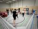 Zwei Boxer im Ring, die einen Sparringskampf durchführen. In den Ringecken sitzen die Trainier der Sportler und schauen zu. Außen um den Ring stehen ander Sportler und Trainer, die ebenfalls zuschauen.