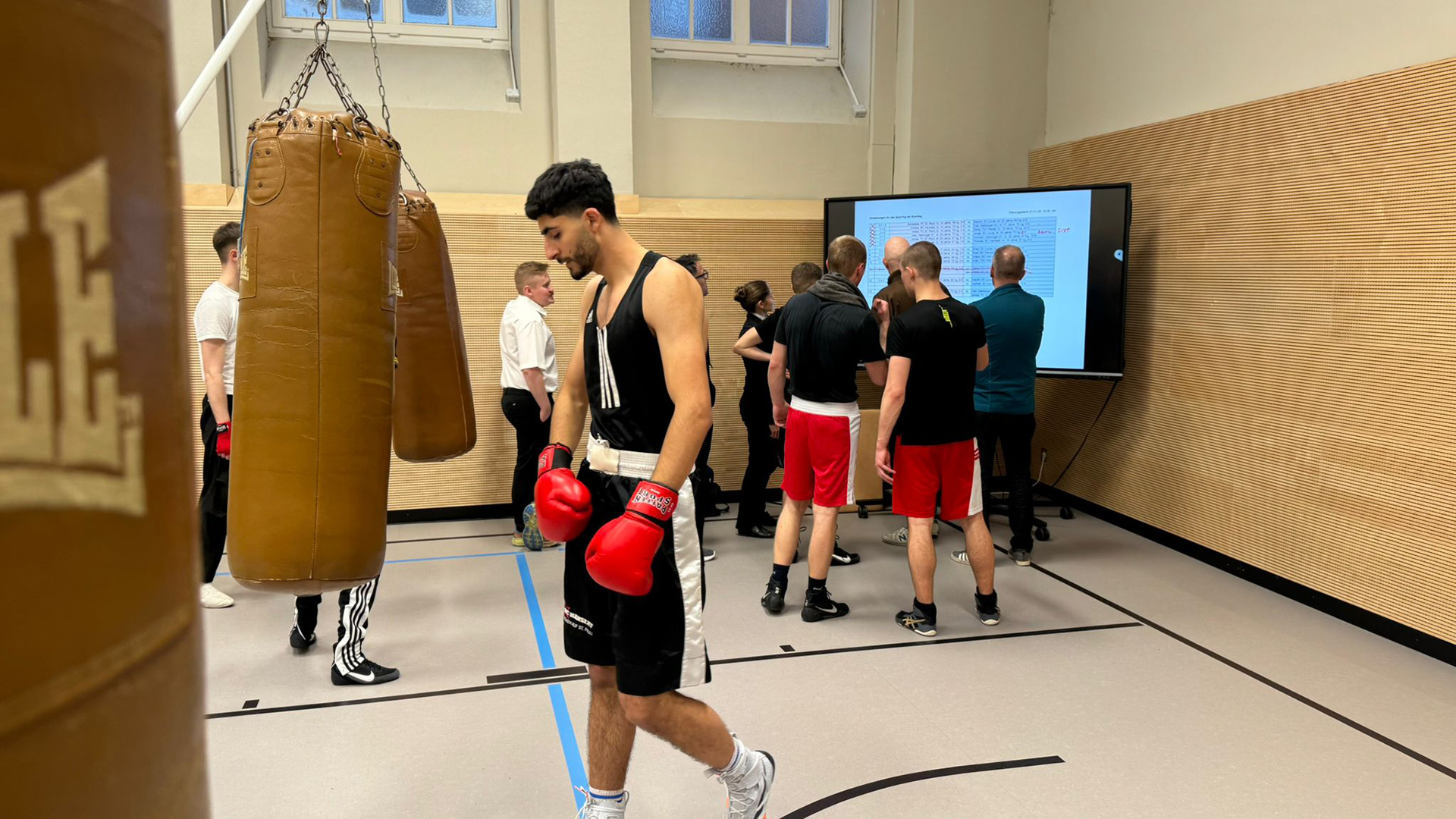 Sportler und Trainer stehen vor einem Screen, auf dem die Ansetzungen der Sparringskämpfe zu sehen sind.